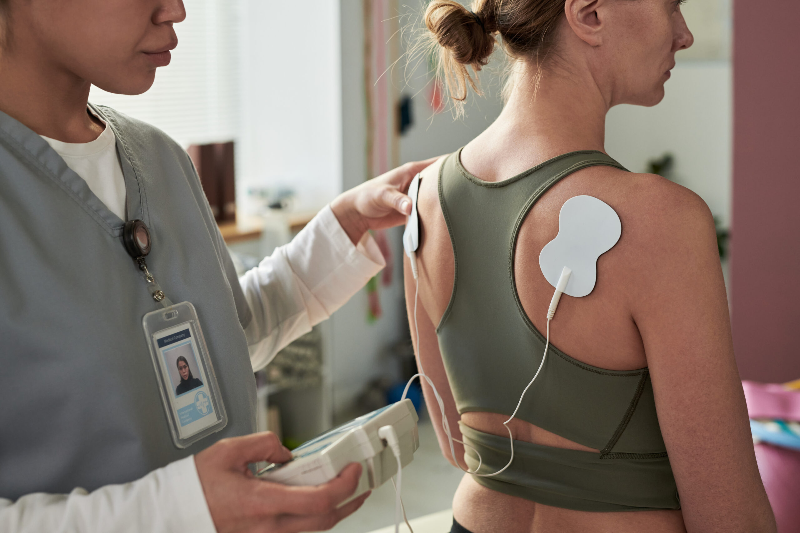 Healthcare professional using TENS unit on patient's back during physical therapy session. Electrode pads applied to patient's back for muscle stimulation and pain relief